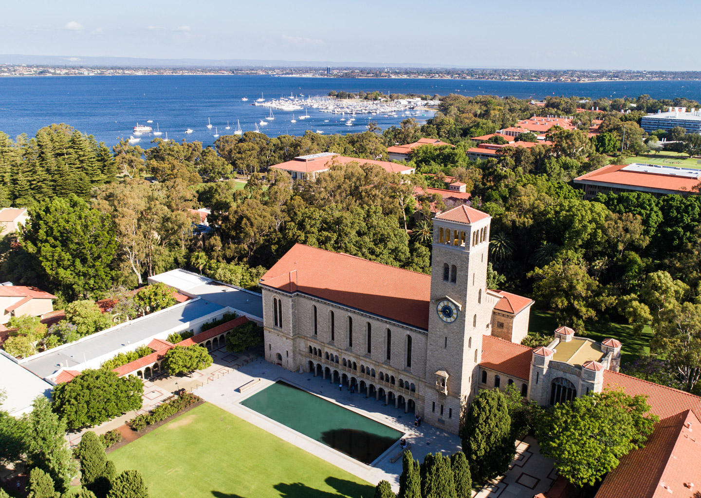 Aerial view of a university campus featuring a historic clock tower, red-roofed buildings, gardens and the river beyond.
