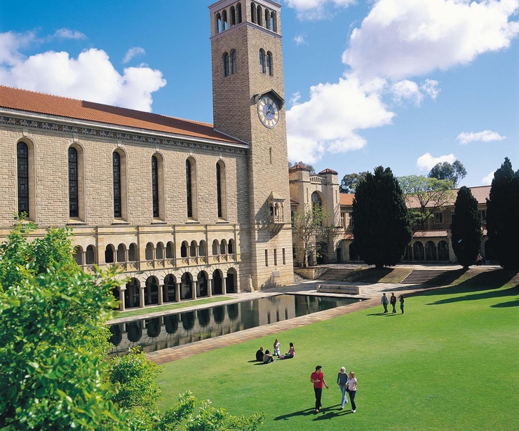Historic sandstone university building with a clock tower, reflecting pool and students on a green lawn.